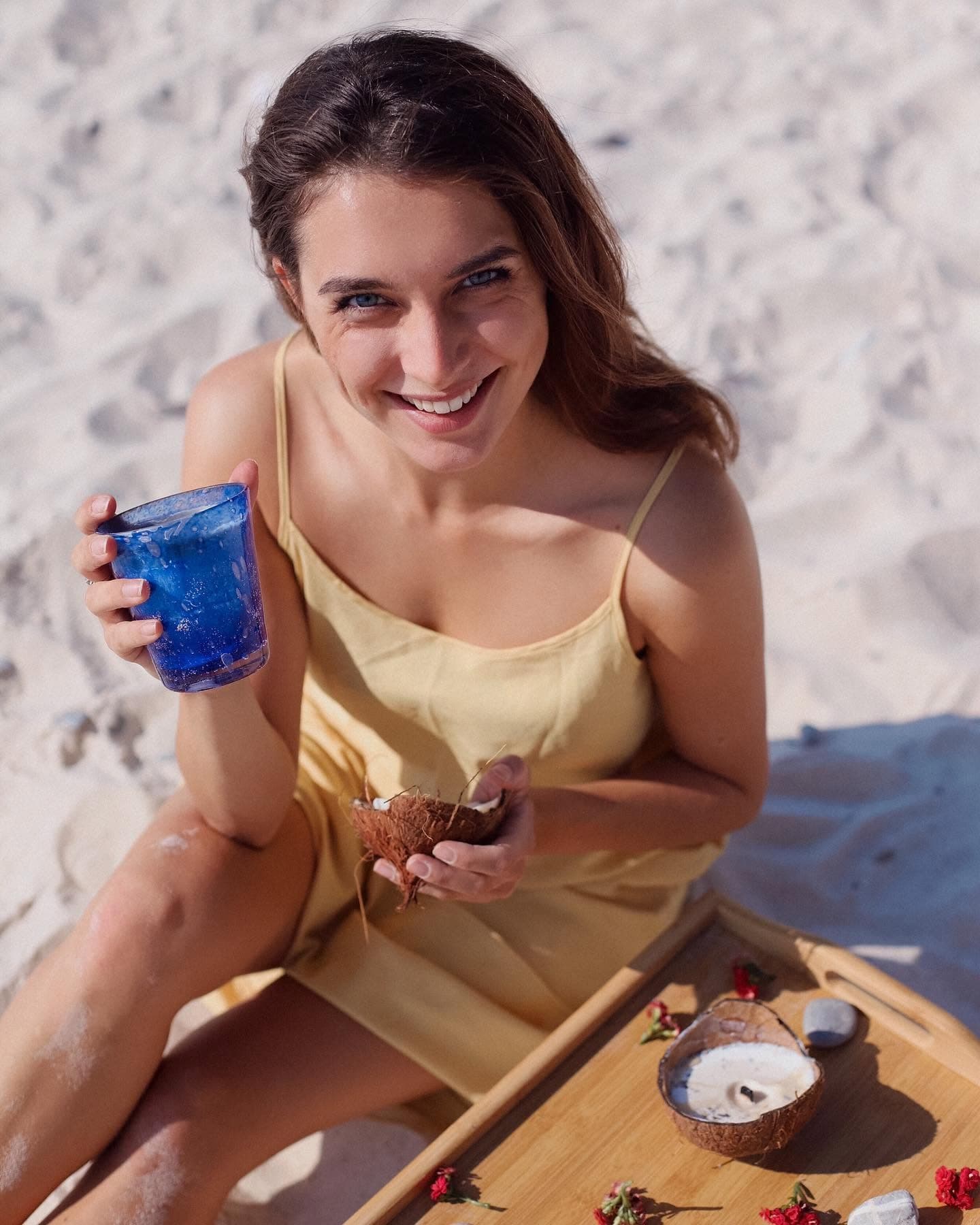 Smiling woman in yellow dress sits on beach holding blue glass and coconut shell.