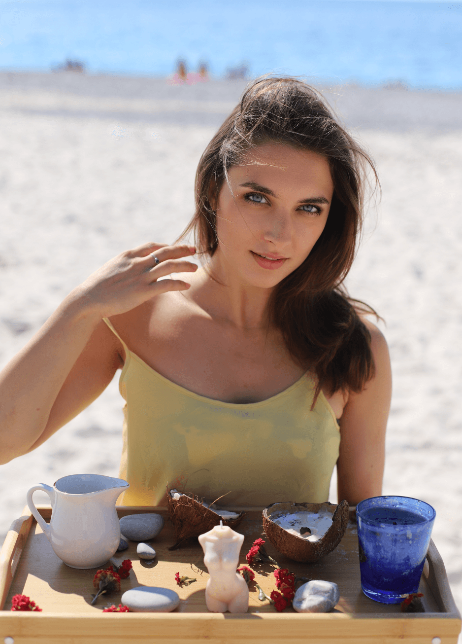 Woman with blue eyes sits behind a tray of spa items on a sunny beach.